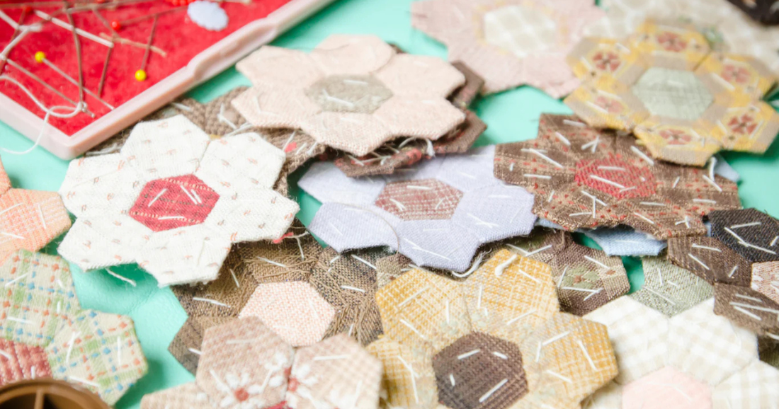 Multiple English paper piecing hexagon flower blocks in neutral, pink and brown fabrics laid out on a teal surface, with a red pin cushion and pins visible in the background