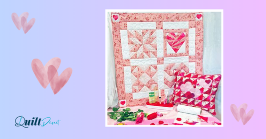 A pink and white heart-themed quilt hanging on a wall alongside a matching heart cushion, sewing notions, and pink roses on a table, set against a pastel purple and blue background with watercolour hearts and the Quilt Direct logo