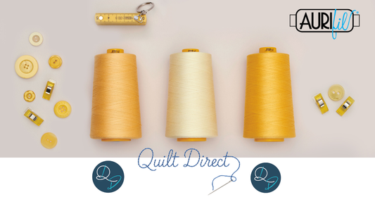 Three large Aurifil thread cones in golden yellow, cream, and deep amber, surrounded by coordinating yellow buttons and fabric clips on a light grey background, with the Quilt Direct logo and Aurifil branding.