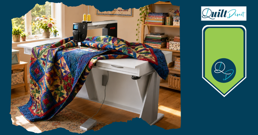 A longarm quilting machine stitching a vibrant patchwork quilt in a bright sewing room, with fabric stacks on shelves in the background – Quilt Direct
