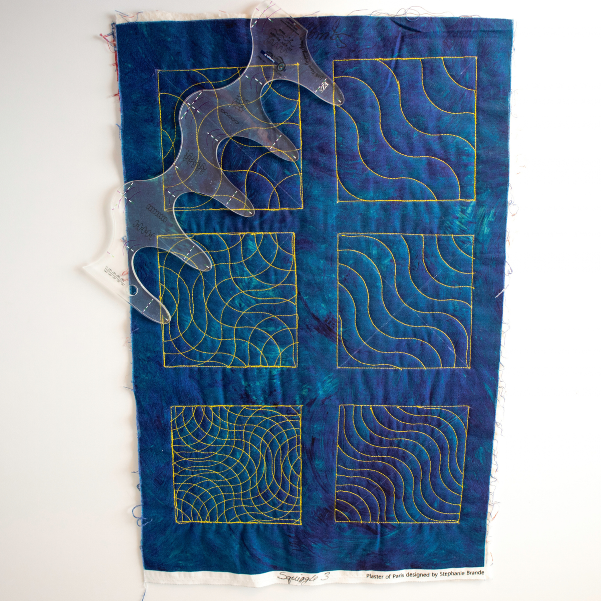 Squiggle Tool 3 on blue fabric showing six quilted blocks with echoed waves and curved crosshatching in yellow thread