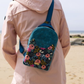 Teal crossbody bag with a floral print front panel, worn by a person on a beach.