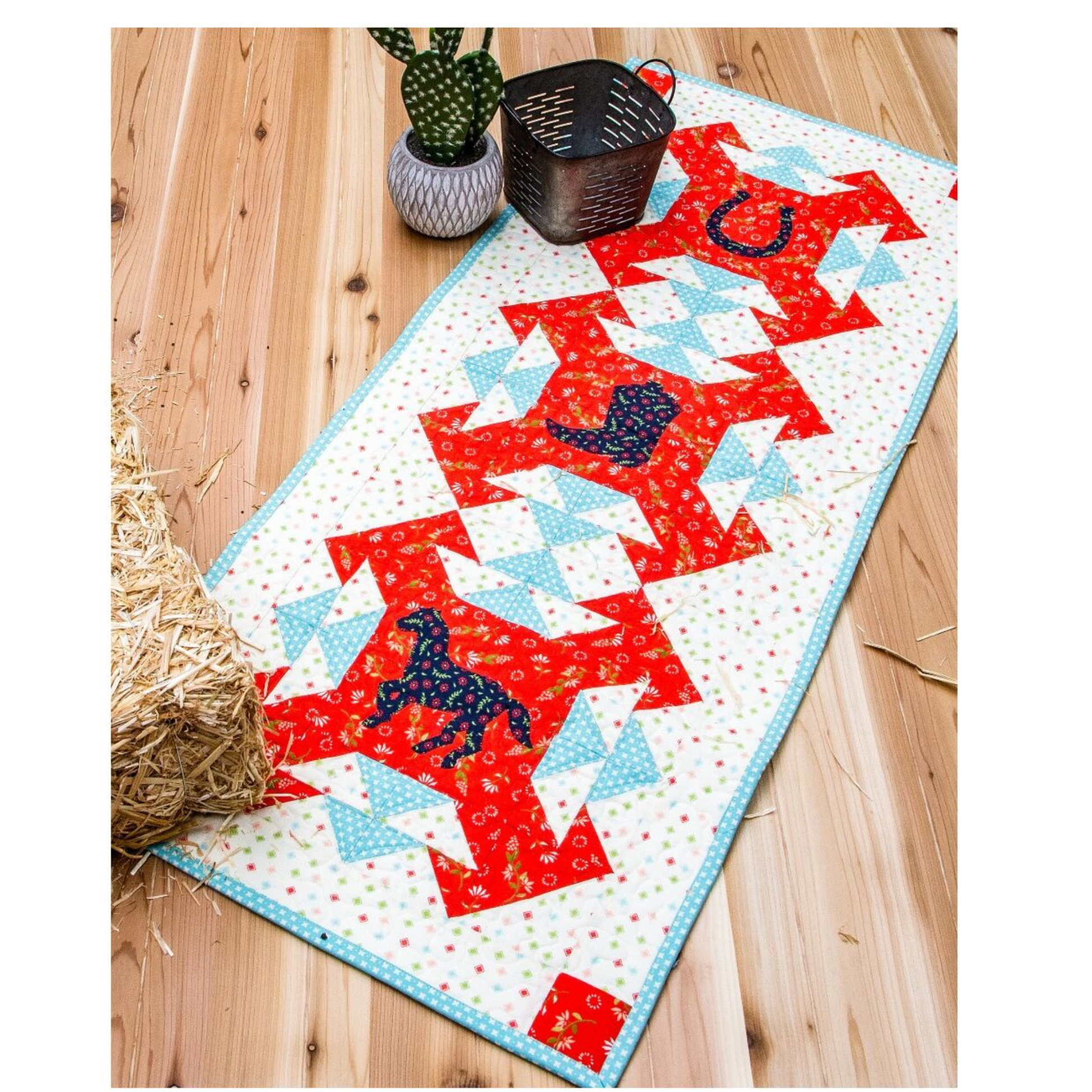 AccuQuilt GO! Western Medley table runner styled on a wood floor with hay, a cactus and a basket