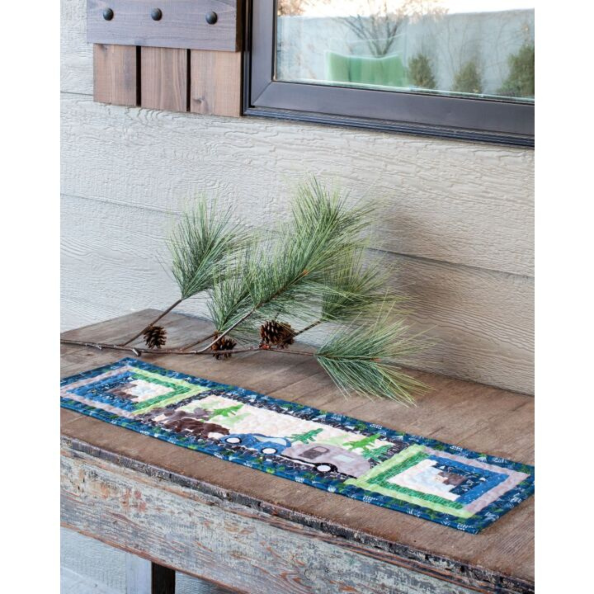 AccuQuilt GO! Camper table runner with a car towing a camper past pine trees, styled with pinecones