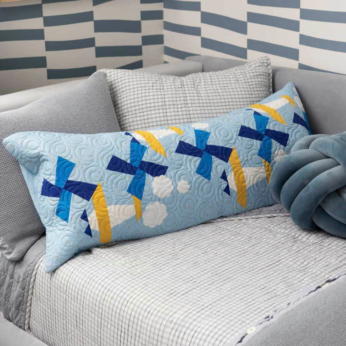 Long quilted bench pillow with blue Airplane blocks, yellow accents and white clouds on a grey sofa