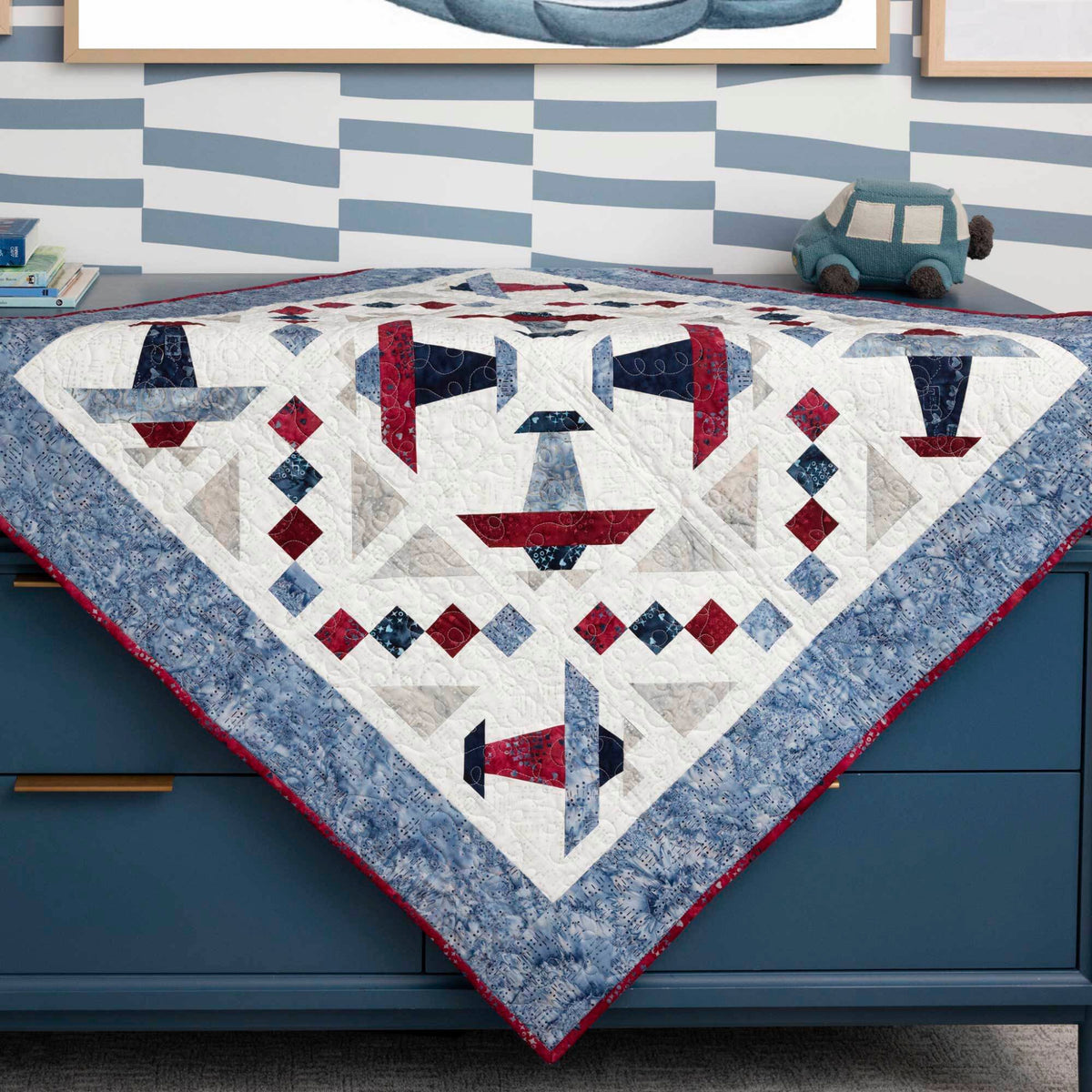 Red, navy and blue Airplane block quilt draped over a blue cabinet, showing its patterned border