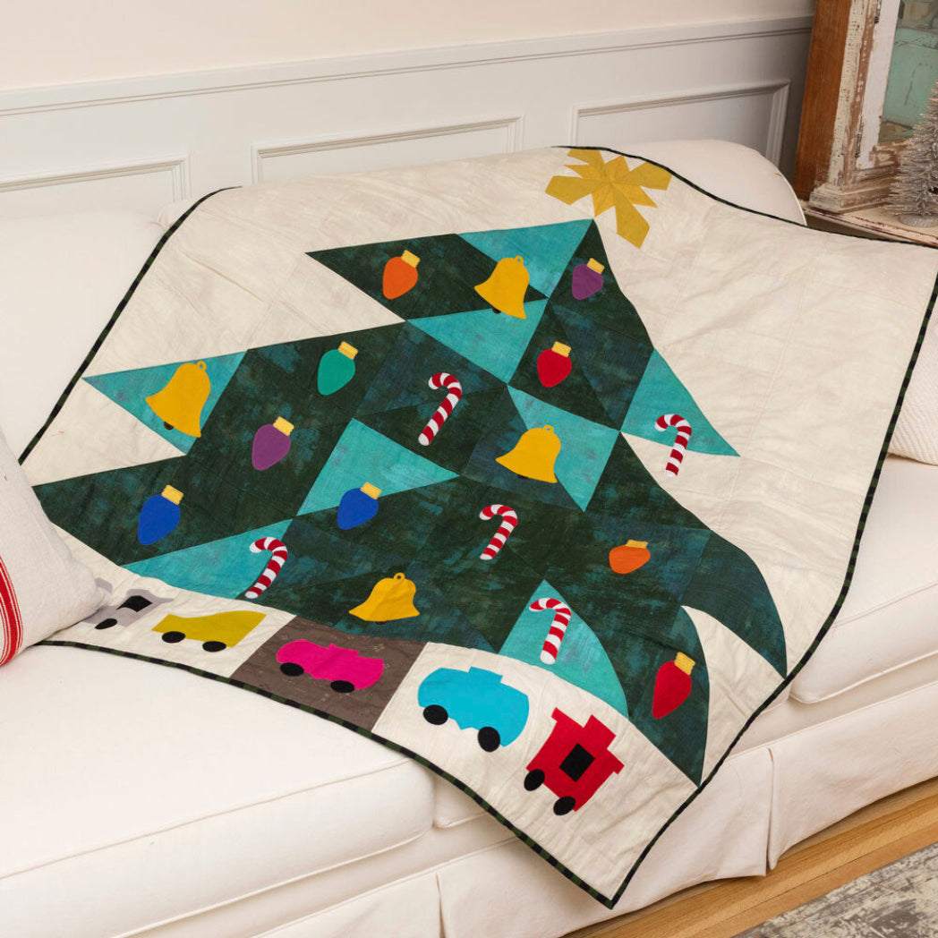 Christmas tree quilt draped over a white sofa, featuring AccuQuilt GO! Jolly Medley appliqué shapes including colourful bells, light bulbs and candy canes on teal and dark green batik, with toy cars and a train border at the base