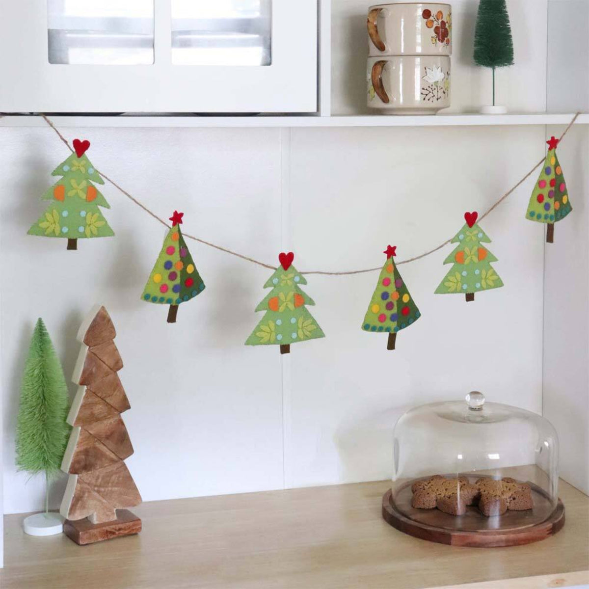 Garland of six decorated fabric Christmas trees made with the AccuQuilt GO! Tinsel die by Sue Spargo, hanging beneath white kitchen cabinets.
