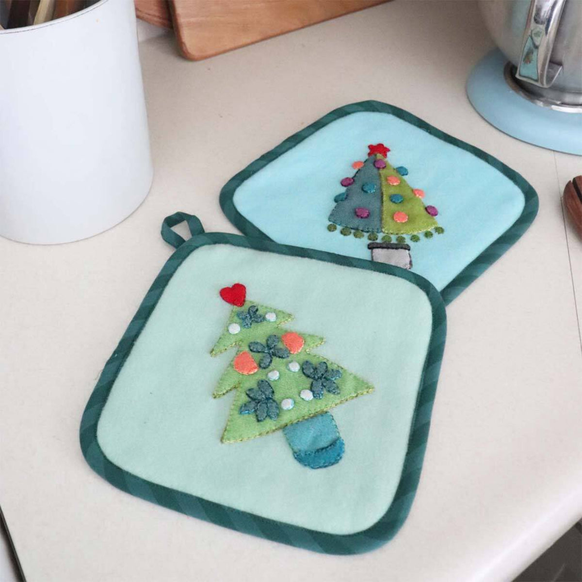 Two pastel potholders with Christmas tree appliqués made using the AccuQuilt GO! Tinsel die by Sue Spargo, displayed on a kitchen worktop.
