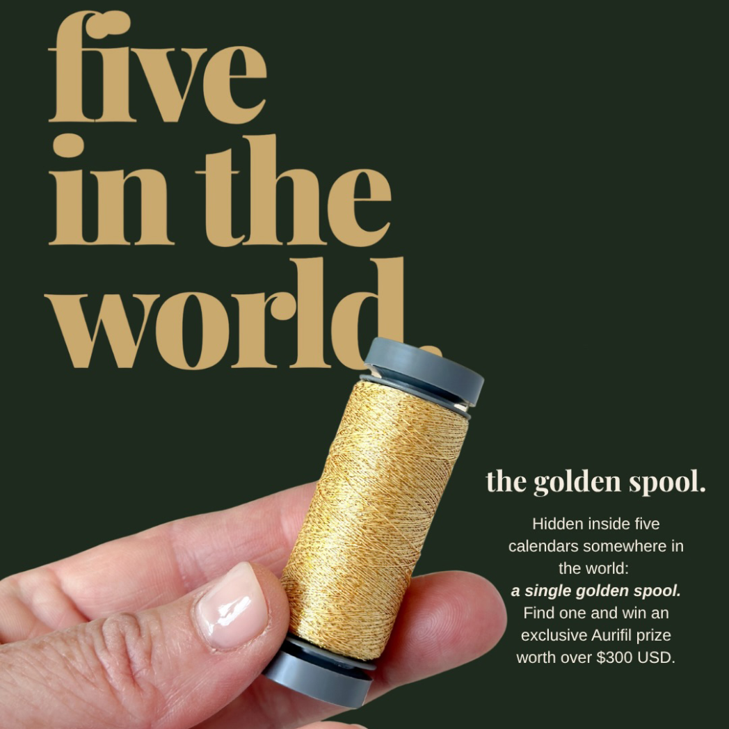 Aurifil Il Salotto 2026 Advent Calendar golden spool, one of five hidden worldwide, held between finger and thumb against a dark green background