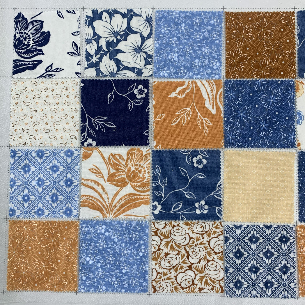 A flat lay of 16 patchwork fabric squares arranged in a 4x4 grid on a cutting mat, featuring floral, ditsy, and geometric prints in navy blue, warm golden tan, soft blue, and cream tones