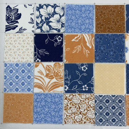 A flat lay of 16 patchwork fabric squares arranged in a 4x4 grid on a cutting mat, featuring floral, ditsy, and geometric prints in navy blue, warm golden tan, soft blue, and cream tones
