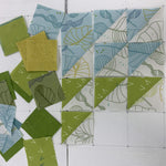 Half-square triangle quilt blocks in progress on a cutting mat, featuring botanical leaf print fabrics in soft blue and olive green tones, with loose fabric squares arranged to the left
