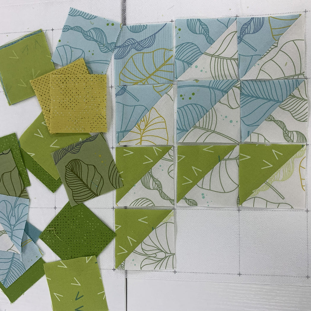 Half-square triangle quilt blocks in progress on a cutting mat, featuring botanical leaf print fabrics in soft blue and olive green tones, with loose fabric squares arranged to the left