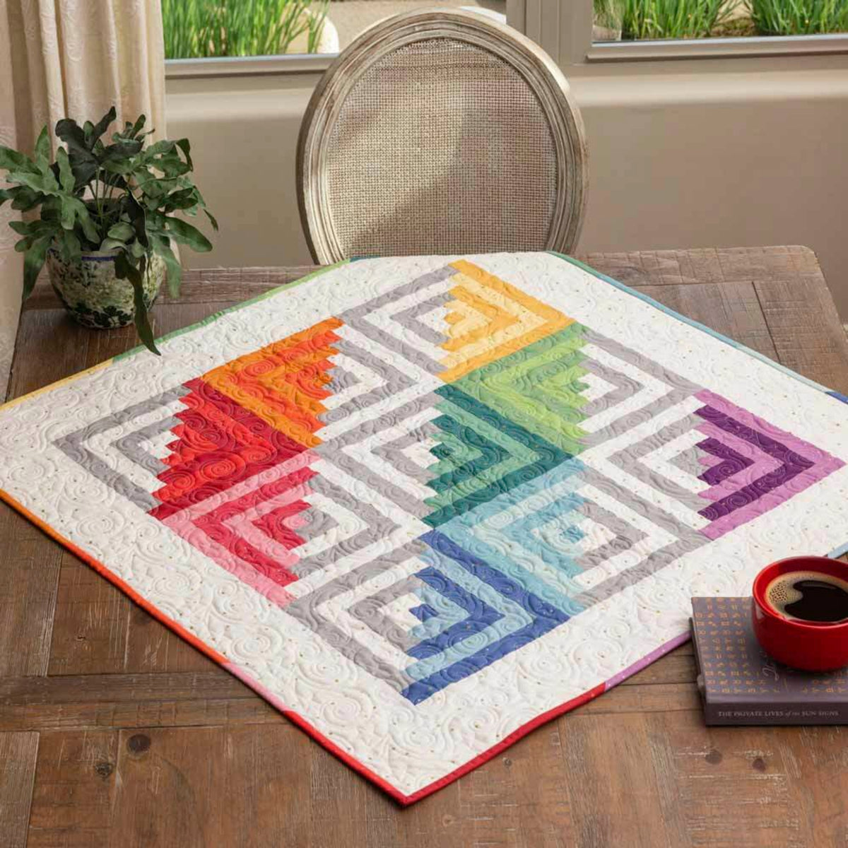 Rainbow-coloured Log Cabin patchwork table topper with a white border laid across a wooden dining table.⠋