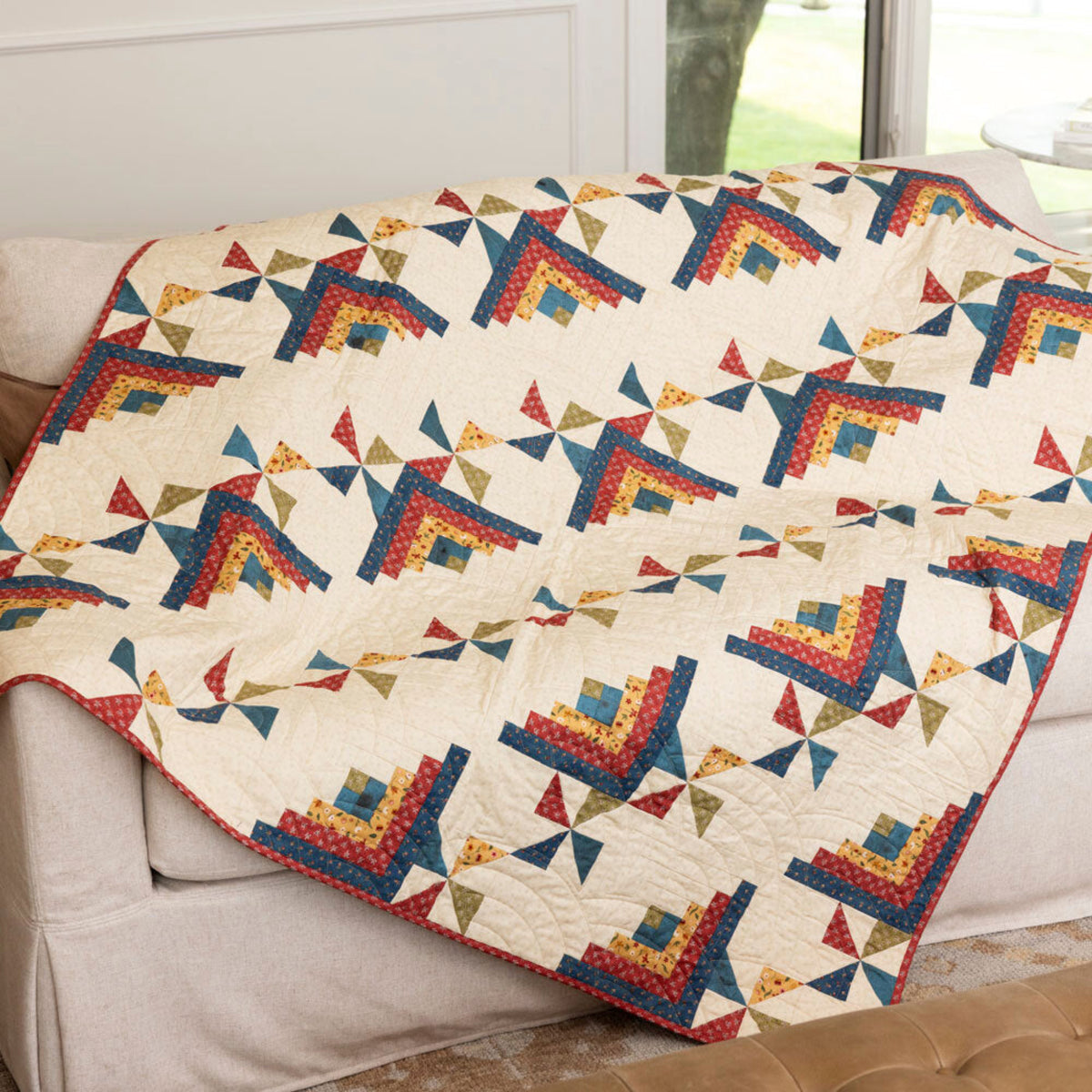 Log Cabin and pinwheel patchwork throw draped over a cream sofa, with red, blue and gold motifs on a cream background.