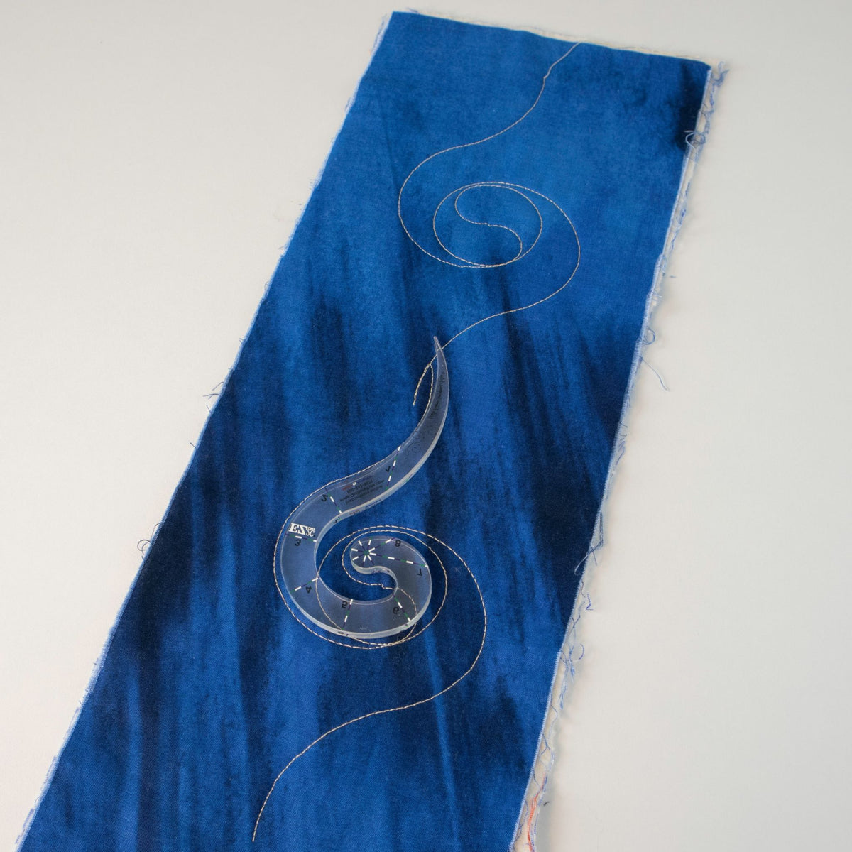 Nautilus Baby Long Tail quilting template on a blue fabric strip with a stitched spiral vine design