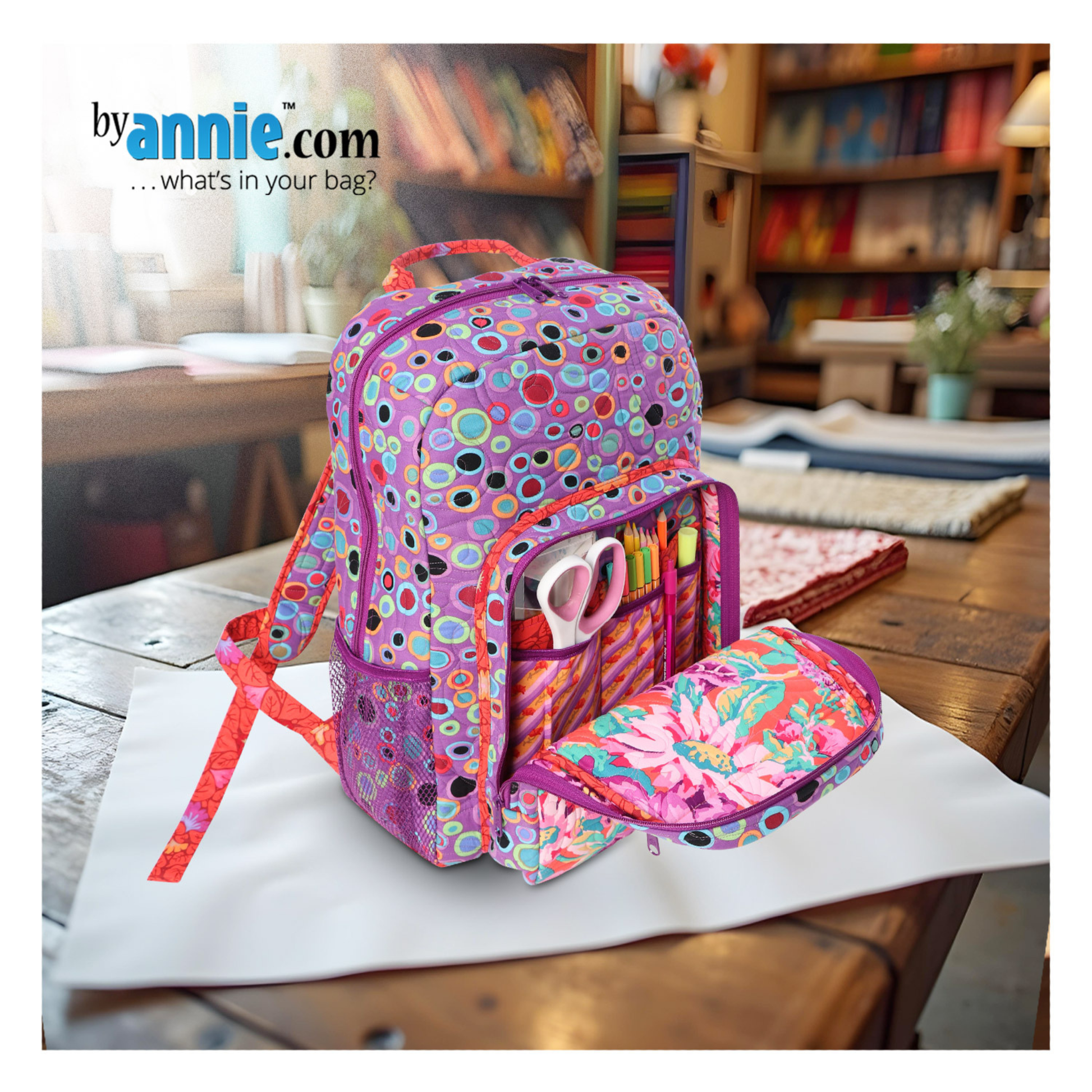 Colorful polka dot backpack with stationery items on a wooden table, byannie.com branding visible.