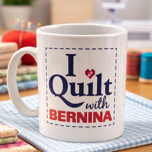 White mug with 'I ❤️ Quilt with BERNINA' text on a checkered tablecloth.