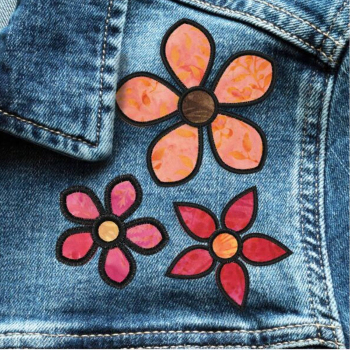 Blue denim embellished with three pink and orange appliqué flowers outlined in black, using AccuQuilt GO! Garden Variety #1 petal and flower centre shapes.