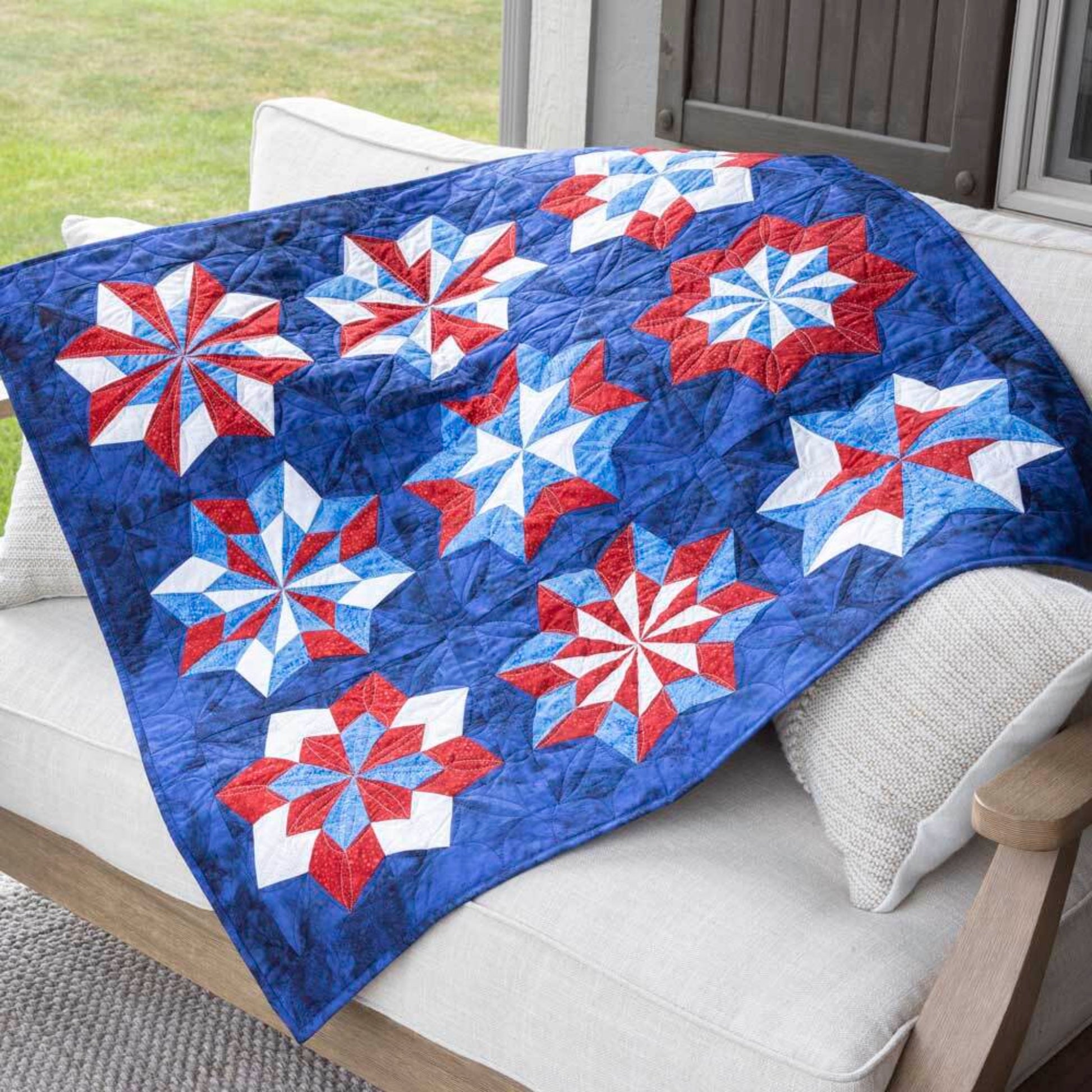 Finished quilt draped over a patio sofa showing repeating double star blocks made with the AccuQuilt GO! Double Star die in red, white and blue fabrics on a navy background, with diamond quilting detail