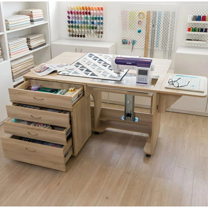 A light wood sewing and quilting table with an attached storage drawer unit on the left, shown in a bright craft room. A purple and white sewing machine sits on the tabletop, which also holds a quilt and a small tray with eyeglasses.
