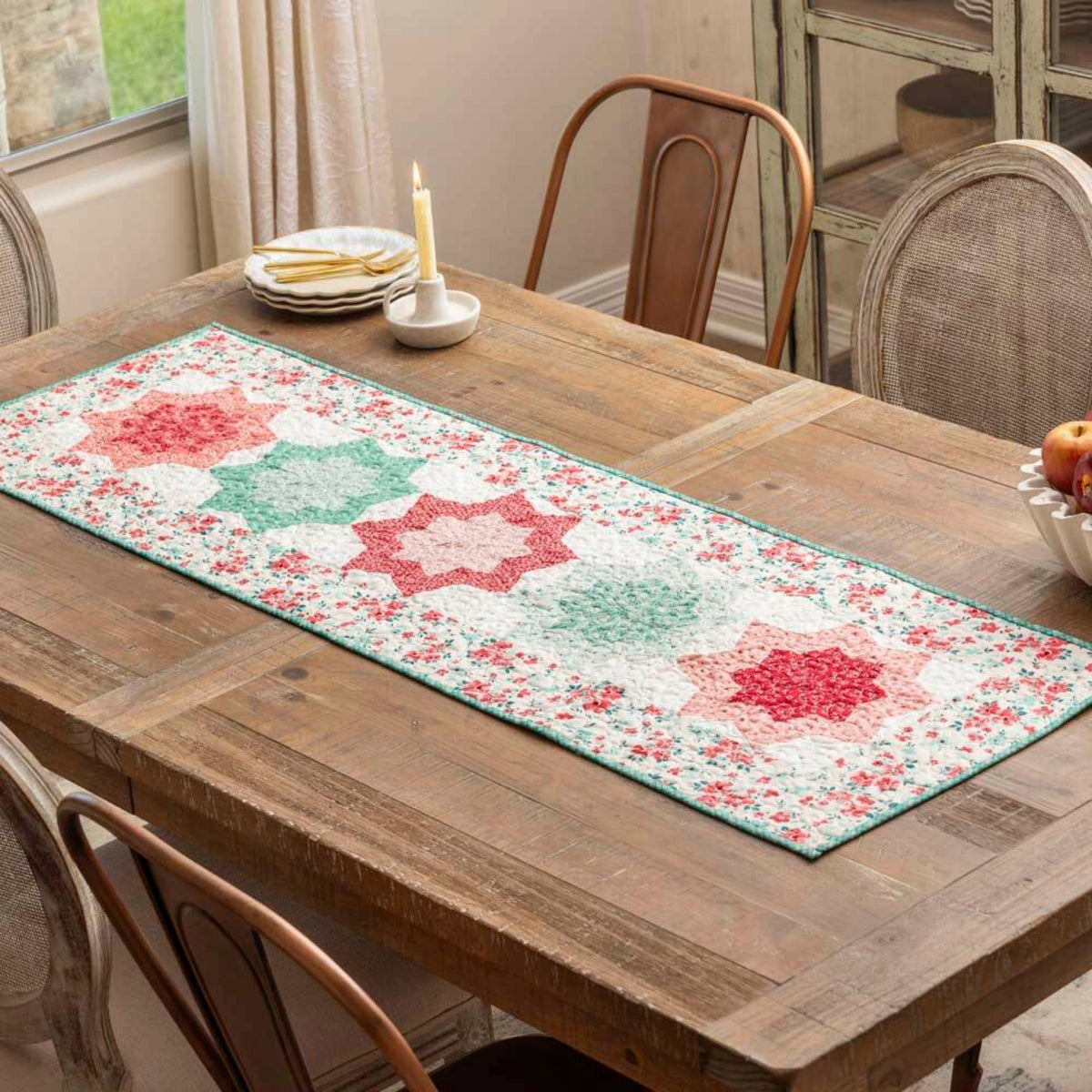 Quilted table runner on a rustic wooden dining table featuring double star blocks made with the AccuQuilt GO! Double Star die in pink, red and sage green floral fabrics on a white ditsy print background